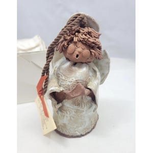 May include: A ceramic figurine of a singing angel with a textured brown clay head and hair, wearing a cream-colored robe with a subtle embossed pattern. The figure features a brown rope loop for hanging and a small rectangular paper tag attached.