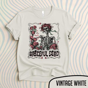 May include: Vintage white t-shirt featuring a skeleton with a floral crown and roses, with the words "Grateful Dead" in a decorative font. The shirt has rolled-up sleeves and a crew neck.