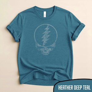 Könnte beinhalten: Ein tealfarbenes T-Shirt mit einem weißen Schädel- und Blitzmotiv in einem Kreis. Die Ärmel sind hochgekrempelt. Der Text "HEATHER DEEP TEAL" befindet sich am unteren Rand des Bildes.
