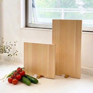 May include: Two rectangular light-colored wooden cutting boards standing upright on small wooden supports. The boards feature a smooth, natural grain finish. Beside the boards, a cluster of fresh red cherry tomatoes and green cucumbers.