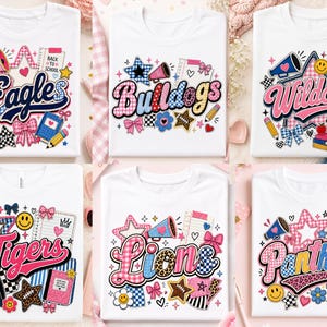以下が含まれることがあります： 学校をテーマにしたカラフルなグラフィックが描かれた6枚の白いTシャツ。各シャツにはEagles、Bulldogs、Wildcats、Tigers、Lions、Panthersといったチーム名が記され、メガホン、星、リボン、ノートなどのイラストが周囲に配置されています。