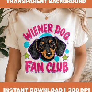 Puede incluir: Una camiseta blanca con un gráfico de la cabeza de un perro salchicha negro y fuego centrado entre el texto rosa "WIENER DOG" y "FAN CLUB". El diseño incluye círculos azul claro y estrellas verde lima rodeando la ilustración.