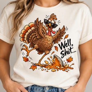 May include: A cream-colored t-shirt featuring a cartoon turkey wearing black sunglasses, looking surprised while standing over a spilled pumpkin pie. The text "Well, Shit..." is printed in black script next to the turkey, surrounded by orange autumn leaves.