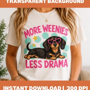 Puede incluir: Una camiseta blanca de manga corta con el gráfico de un perro salchicha negro y fuego que lleva gafas de sol rosas en forma de corazón. El texto "MORE WEENIES" y "LESS DRAMA" aparece en rosa sobre y bajo el perro, con puntos azul claro.