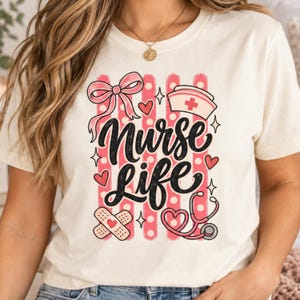 Puede incluir: Una camiseta de color crema con un diseño gráfico que incluye el texto "Nurse Life" en letra cursiva negra. El diseño presenta rayas verticales rosas, lunares blancos, un lazo rosa, una cofia de enfermera, un estetoscopio en forma de corazón y tiritas.