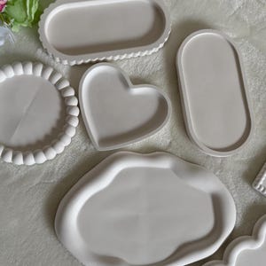 May include: A collection of white decorative trays in various shapes including a round scalloped tray, an oval tray, a heart-shaped tray, an irregular organic-shaped tray, and a rectangular tray with a bow detail, all featuring smooth surfaces.