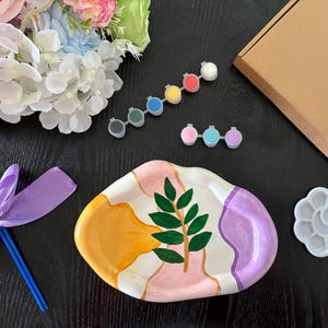 May include: An oval ceramic tray with a hand-painted green leaf design, yellow, pink, and purple abstract shapes, and gold accents. Includes a paintbrush with a purple ribbon, two strips of paint pots, a flower-shaped palette, and a cardboard box.