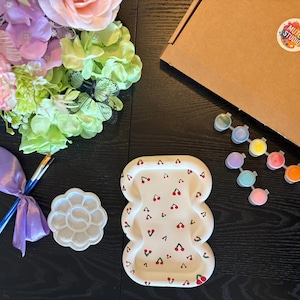 May include: A craft kit featuring a white rectangular tray with a red cherry pattern, a flower-shaped paint palette, two paintbrushes tied with a purple ribbon, a strip of eight small paint pots, and a cardboard box labelled Muted Studio.