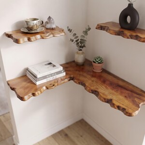 Peut inclure: Trois étagères flottantes en forme de L en bois naturel avec des bords bruts. Les étagères sont fixées dans un angle et supportent une tasse et une soucoupe en céramique, un cristal, une pile de livres, un vase avec eucalyptus et une plante.