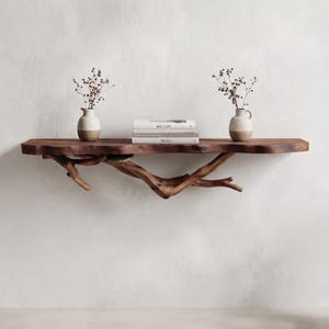 May include: A wall-mounted shelf made from a thick, dark brown live-edge wood slab supported by a natural, gnarled tree branch. Two small, two-toned ceramic vases containing dried floral stems and a stack of three books sit on the shelf surface.