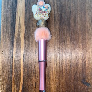 May include: A pink metal pen featuring a decorative topper with a peach-colored fuzzy pom-pom, a metallic gold bead, and a white butterfly-shaped bead adorned with a floral pattern and the word Gucci in gold script lettering.