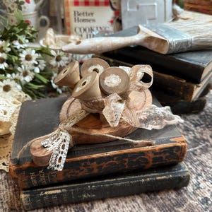 May include: Three small wooden spools are arranged on a circular wooden base, tied together with a piece of delicate, off-white lace ribbon. The spools have a natural light brown finish and are positioned to show their cylindrical shape and center holes.