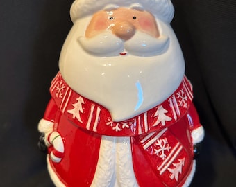 Puede incluir: Una figura de cerámica de Santa Claus con un acabado brillante en rojo y blanco. El personaje lleva un gorro rojo con un pompón blanco y una bufanda roja decorada con patrones de copos de nieve y árboles blancos. Sostiene un bastón de caramelo.