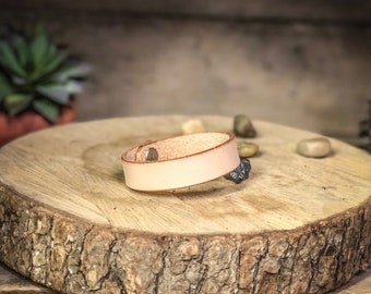 May include: A light brown leather bracelet with a silver buckle on a wooden surface.