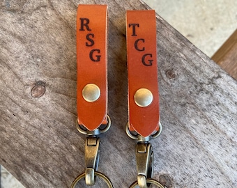 Pode incluir: Dois chaveiros de couro castanho com argolas de chave de latão e fechos de metal. Os chaveiros são personalizados com as iniciais "RSG" e "TCG" em preto.
