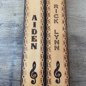 Pu&ograve; includere: Due cinturini per chitarra in pelle color cuoio con cuciture decorative nere e un design a chiave di violino. I cinturini sono personalizzati con i nomi "Aiden" e "Rick Lynn".