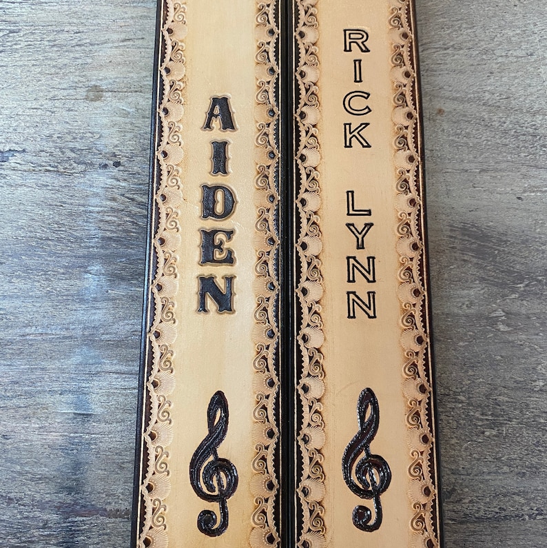 Pu&ograve; includere: Due cinturini per chitarra in pelle color cuoio con cuciture decorative nere e un design a chiave di violino. I cinturini sono personalizzati con i nomi "Aiden" e "Rick Lynn".