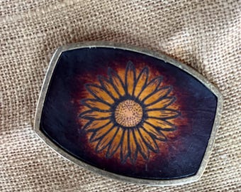 May include: A rectangular belt buckle with a silver-coloured frame. The buckle features a sunflower design with yellow petals and a brown centre on a dark brown background.