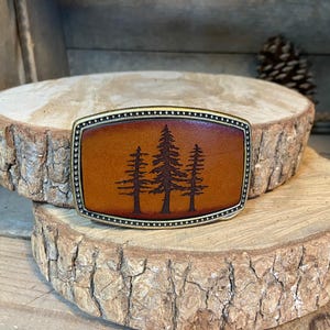 A leather belt buckle with a tree silhouette design is displayed on a wooden surface.