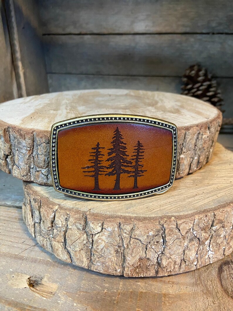A leather belt buckle with a tree silhouette design is displayed on a wooden surface.