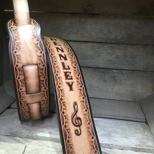 Pu&ograve; includere: Un cinturino per chitarra in pelle marrone chiaro con un design decorativo e il nome "ZENNLEY" e una chiave di violino incisa.