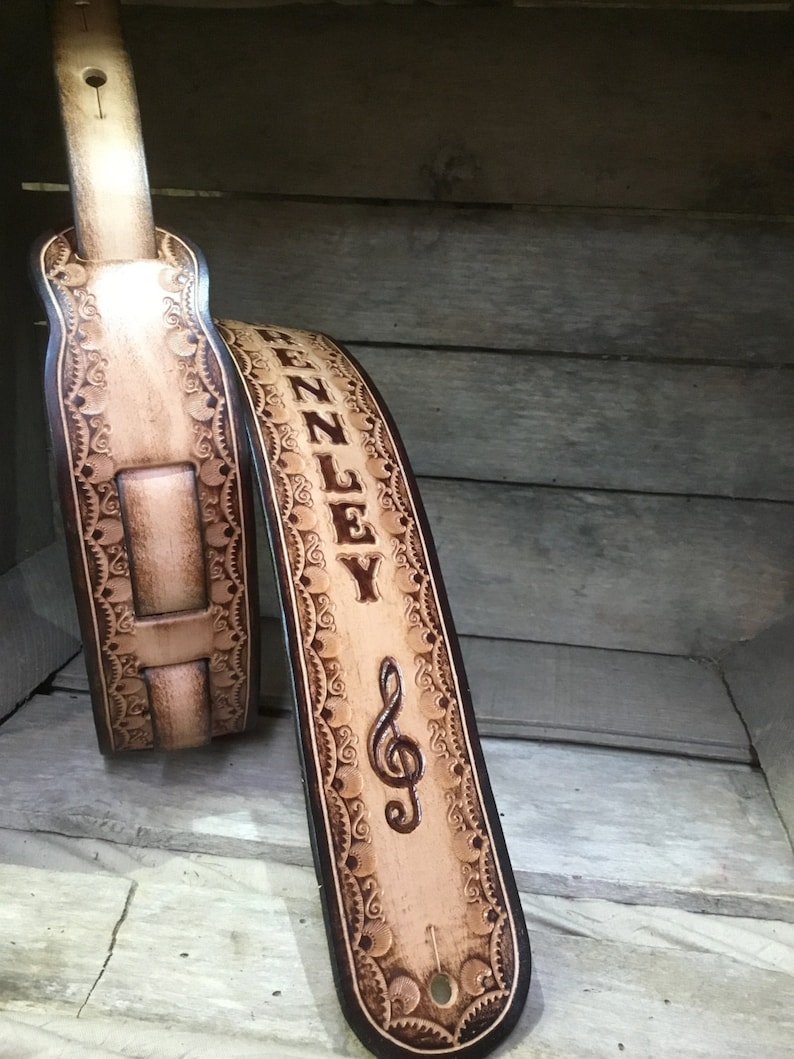 Pu&ograve; includere: Un cinturino per chitarra in pelle marrone chiaro con un design decorativo e il nome "ZENNLEY" e una chiave di violino incisa.