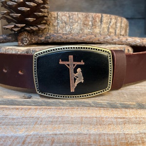 May include: A brown leather belt with a brass buckle featuring a silhouette of a lineman working on a power pole.
