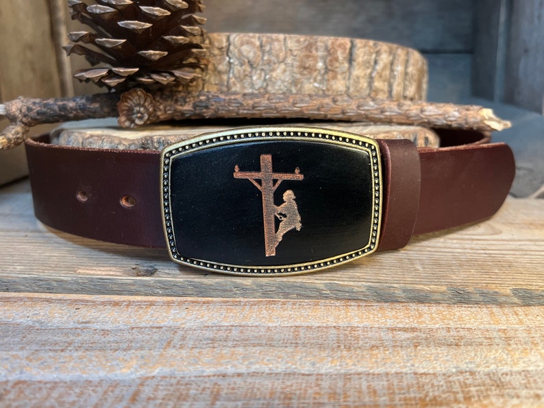May include: A brown leather belt with a brass buckle featuring a silhouette of a lineman working on a power pole.