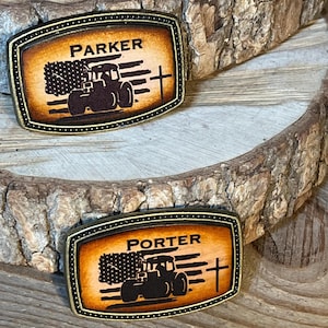 May include: Two personalized leather belt buckles with a tractor, American flag, and cross design. The top buckle says "PARKER" and the bottom buckle says "PORTER".