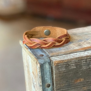 May include: A braided leather bracelet with a brown and tan color scheme. The bracelet has a snap closure.