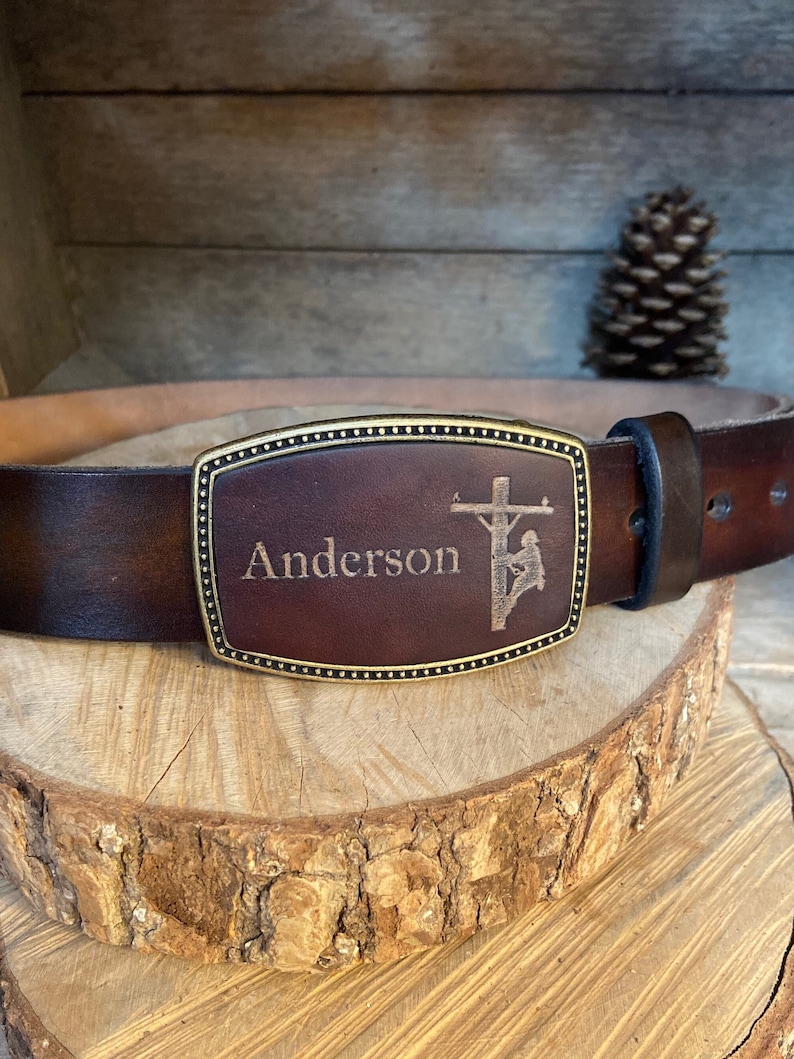 May include: A brown leather belt with a brass buckle featuring an engraved image of a lineman working on a power line. The buckle is personalized with the name "Anderson".