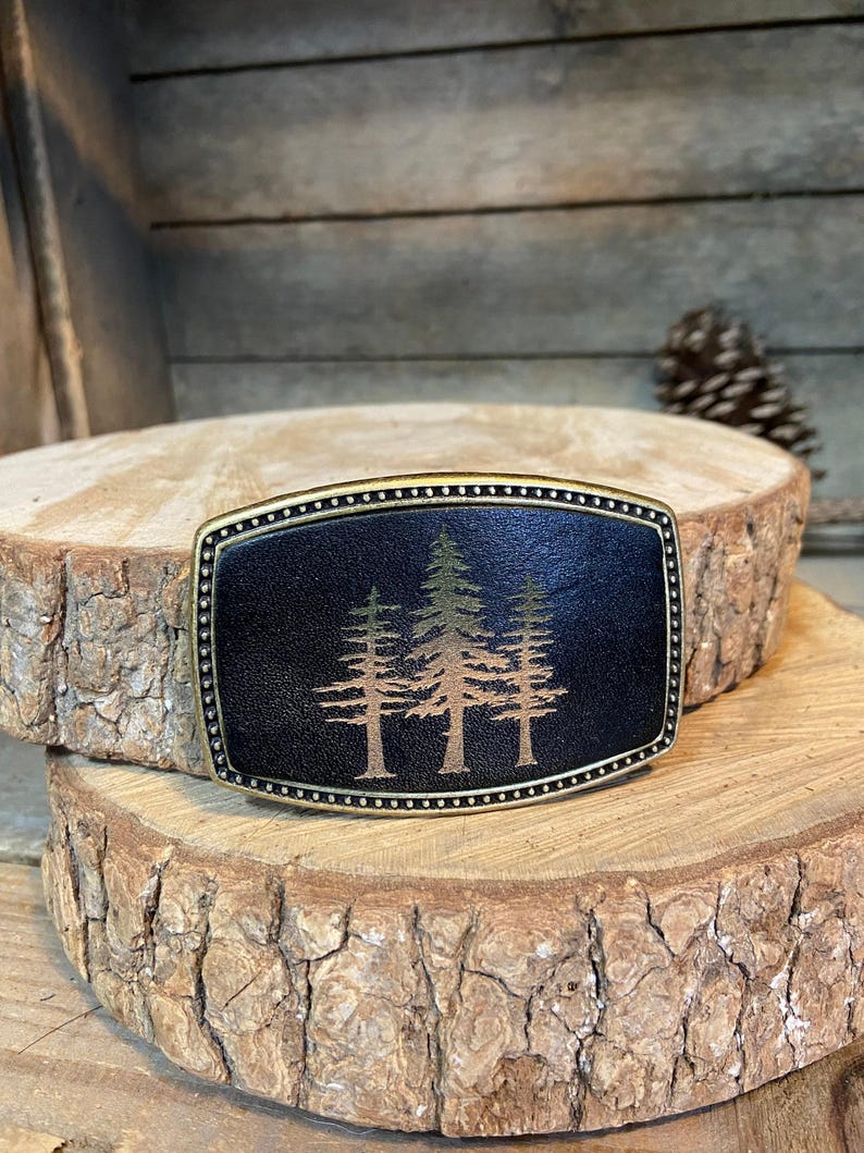 A black leather belt buckle with a tree design on it, placed on a wooden surface.