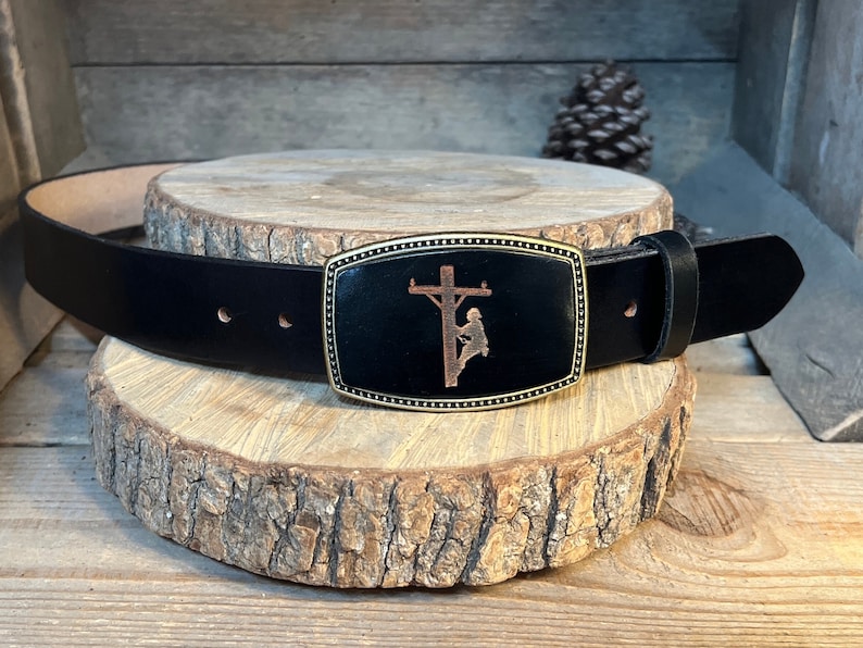 May include: A black leather belt with a gold-toned buckle featuring a silhouette of a lineman working on a power pole.