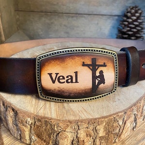 May include: A brown leather belt with a brass buckle. The buckle is engraved with a silhouette of a lineman working on a power pole and the name "Veal".