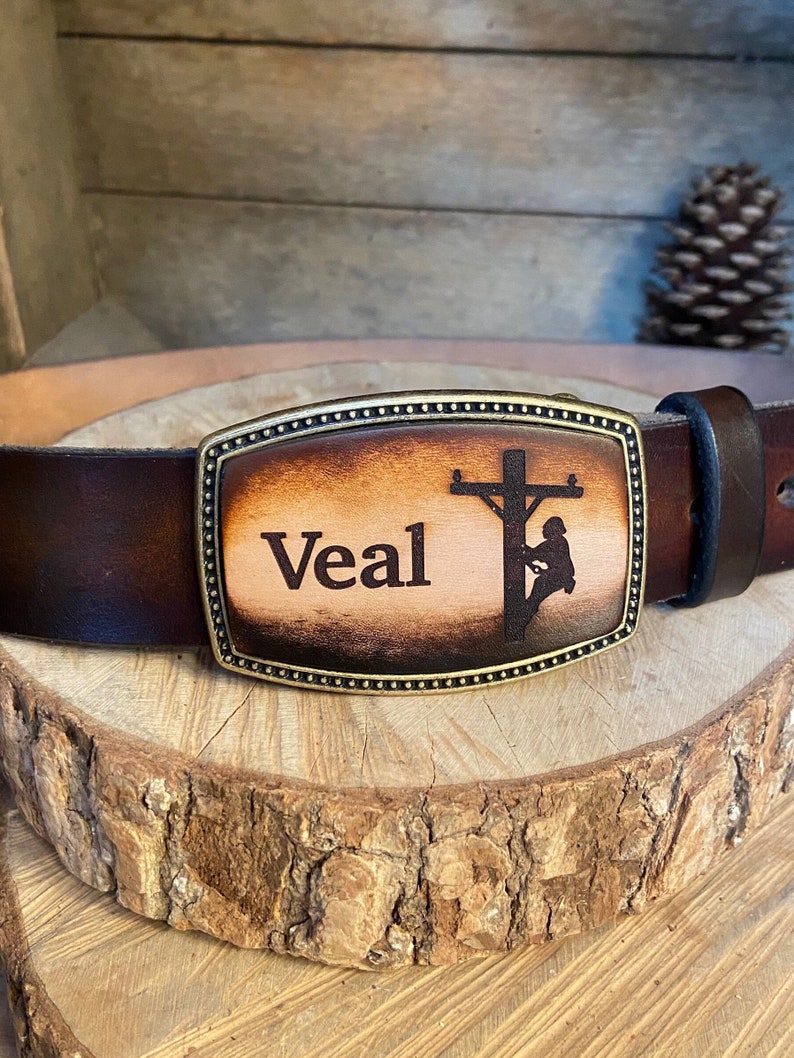 May include: A brown leather belt with a brass buckle. The buckle is engraved with a silhouette of a lineman working on a power pole and the name "Veal".