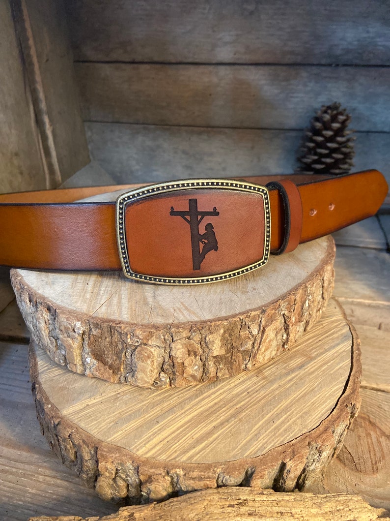 May include: A brown leather belt with a brass buckle featuring a silhouette of a lineman working on a power pole.
