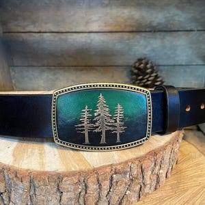 A black leather belt with a decorative metal buckle featuring a tree design, placed on a wooden surface.