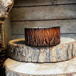May include: A brown leather cuff bracelet with a design of tall grass and wheat stalks.