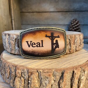 May include: A brown leather belt buckle with a brass frame. The buckle features a silhouette of a lineman working on a power pole. The name "Veal" is engraved on the buckle.