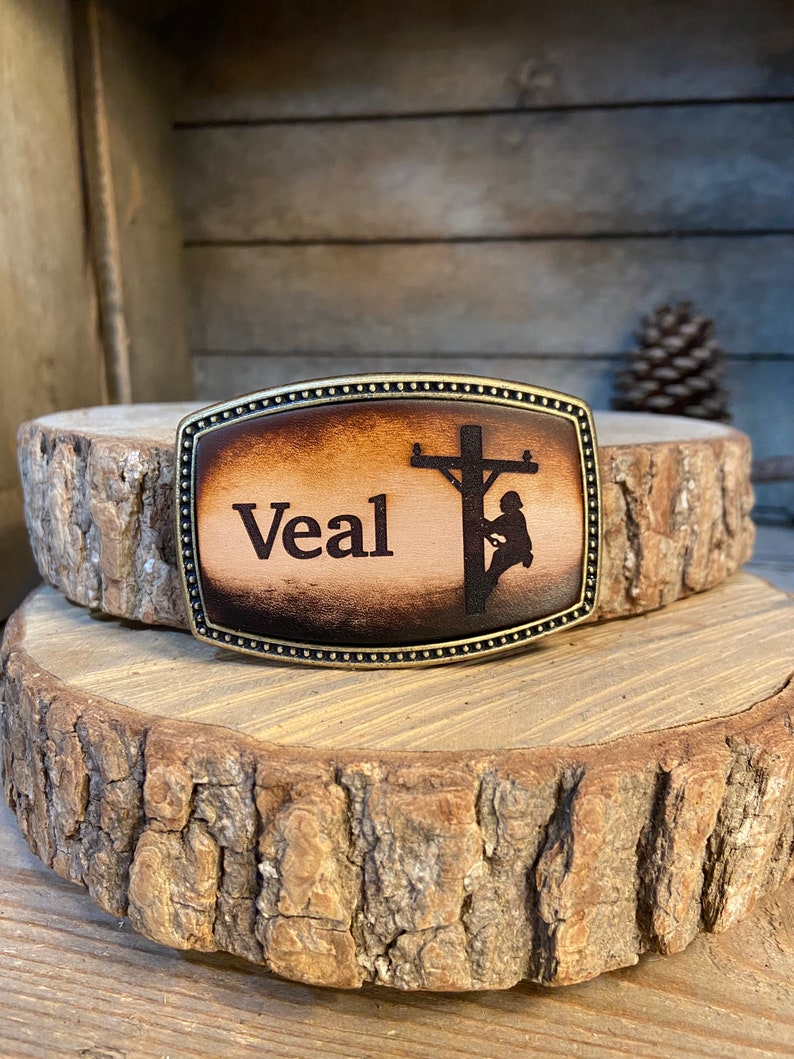 May include: A brown leather belt buckle with a brass frame. The buckle features a silhouette of a lineman working on a power pole. The name "Veal" is engraved on the buckle.