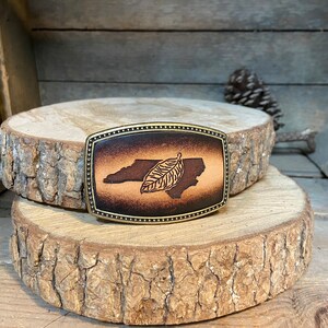 Puede incluir: Una hebilla de cintur&oacute;n de cuero marr&oacute;n con un dise&ntilde;o del estado de Carolina del Norte y una hoja. La hebilla es de color lat&oacute;n y tiene un borde decorativo.