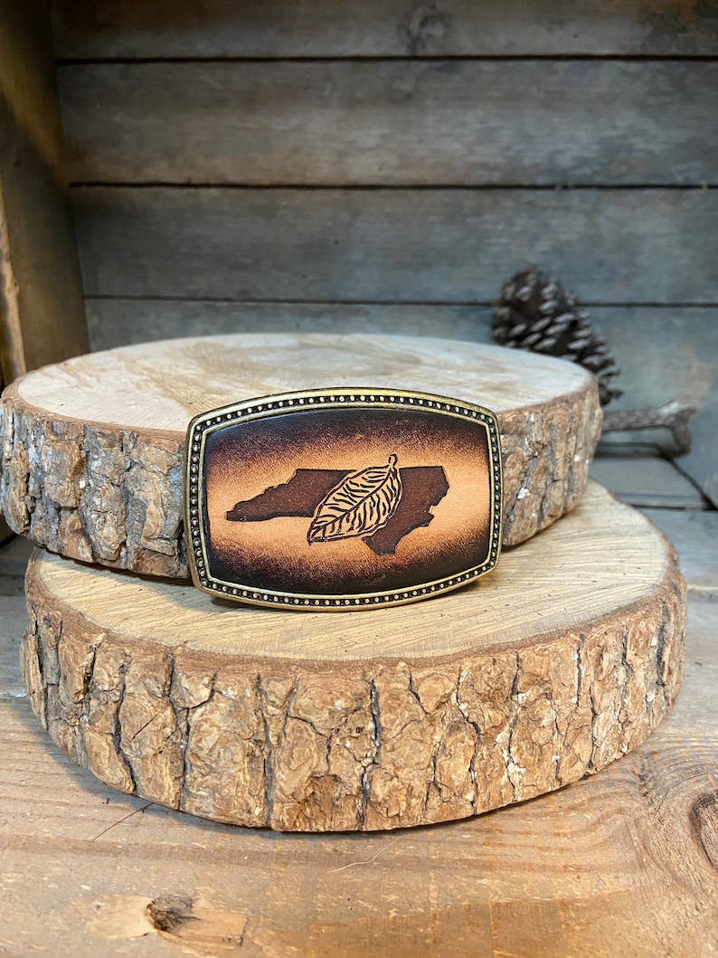 Puede incluir: Una hebilla de cintur&oacute;n de cuero marr&oacute;n con un dise&ntilde;o del estado de Carolina del Norte y una hoja. La hebilla es de color lat&oacute;n y tiene un borde decorativo.