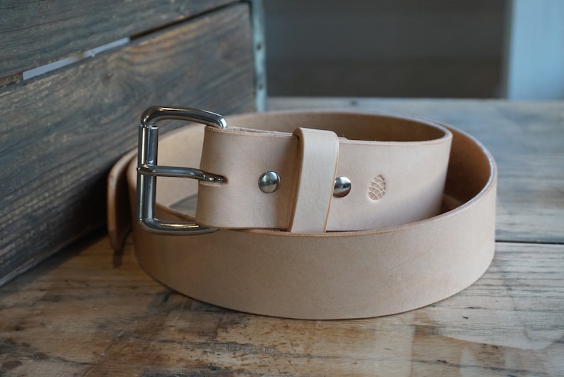 K&ouml;nnte beinhalten: Ein hellbrauner Lederg&uuml;rtel mit einer silbernen Schnalle. Der G&uuml;rtel hat ein kleines, gepr&auml;gtes Design auf dem Leder.