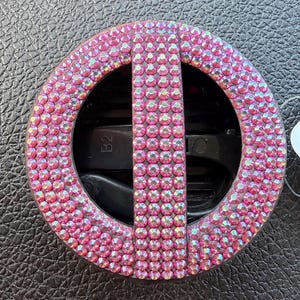 May include: A circular hair clip featuring a black plastic base covered in small, iridescent pink rhinestones. The design includes a vertical bar across the center, and the interior mechanism is marked with the alphanumeric text B2.