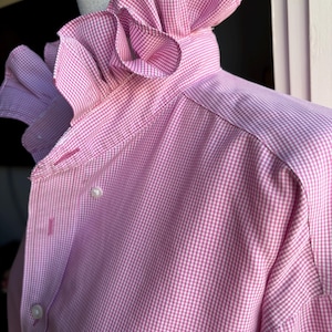 May include: A long-sleeved button-down shirt in a pink and white gingham pattern. The shirt features a high, standing collar with a prominent, ruffled trim that extends around the neckline. The front placket includes small white buttons.