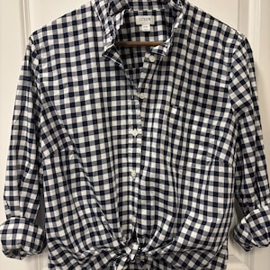May include: A navy blue and white gingham button-down shirt with a ruffled collar, long sleeves with rolled cuffs, and a front tie hem. The shirt features a front button placket, a chest pocket, and a J.Crew label indicating size XXL.
