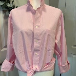 May include: A light pink long-sleeve button-down shirt featuring a ruffled stand-up collar, a single chest pocket, and cuffs that are folded upward. The shirt is tied at the front hem and displays a series of white buttons down the center placket.
