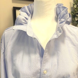 May include: A light blue and white gingham button-down shirt featuring a prominent ruffled collar. The shirt is displayed on a mannequin and includes a single chest pocket on the lower right side, with visible white buttons down the front.