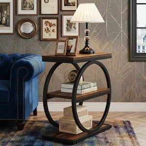 May include: A rectangular side table with a dark wood finish and a black metal frame featuring two large circular side supports. The table has three tiers, holding a table lamp with a white pleated shade, two books, and two stacked decorative boxes.