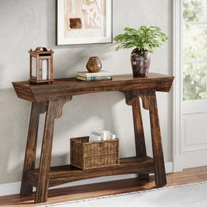 May include: A dark brown wooden console table with a rectangular top and a lower shelf. The table features angled legs and a smooth finish. On the top surface sits a wooden lantern, a small brown vase, and two stacked books. A woven basket sits below.